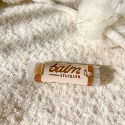 A tube of Balm Standard lip balm lies on a soft, textured white blanket with a fluffy white pom-pom nearby. The label is cream-colored with brown text and accents.