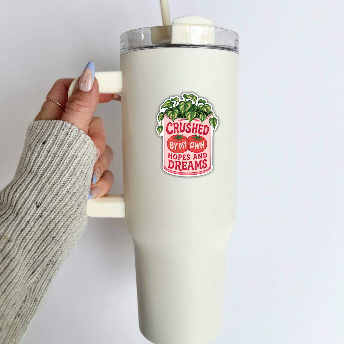 A hand wearing a gray ribbed knit sweater holds a cream-colored insulated travel tumbler with a clear lid and straw. The tumbler features a sticker that reads 'CRUSHED BY MY OWN HOPES AND DREAMS' with a green leafy design.