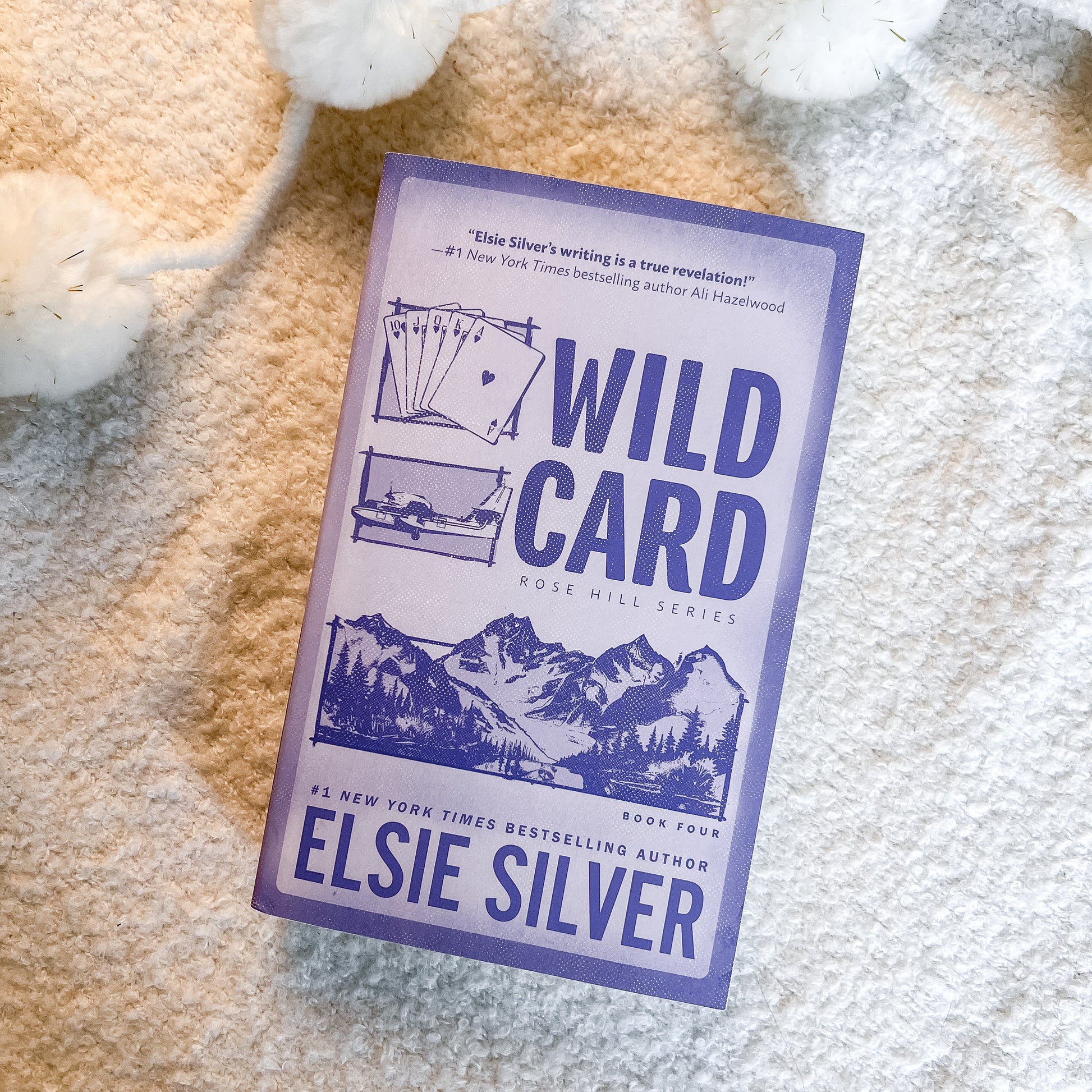 The book 'Wild Card: Rose Hill Series' by Elsie Silver, featuring a lavender and white cover with blue illustrations, photographed flat on a textured cream-colored blanket.