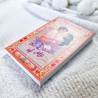 A paperback book titled 'You'll Be the Death of Me' by Holly Michelle, featuring a pink and orange vintage-style cover with an illustration of a couple, resting on a white textured blanket.