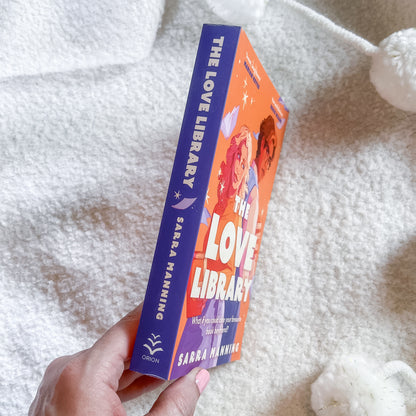A hand holds a book titled 'The Love Library' by Sarah Manning, showing the orange and purple cover and spine, set against a soft white textured blanket background.