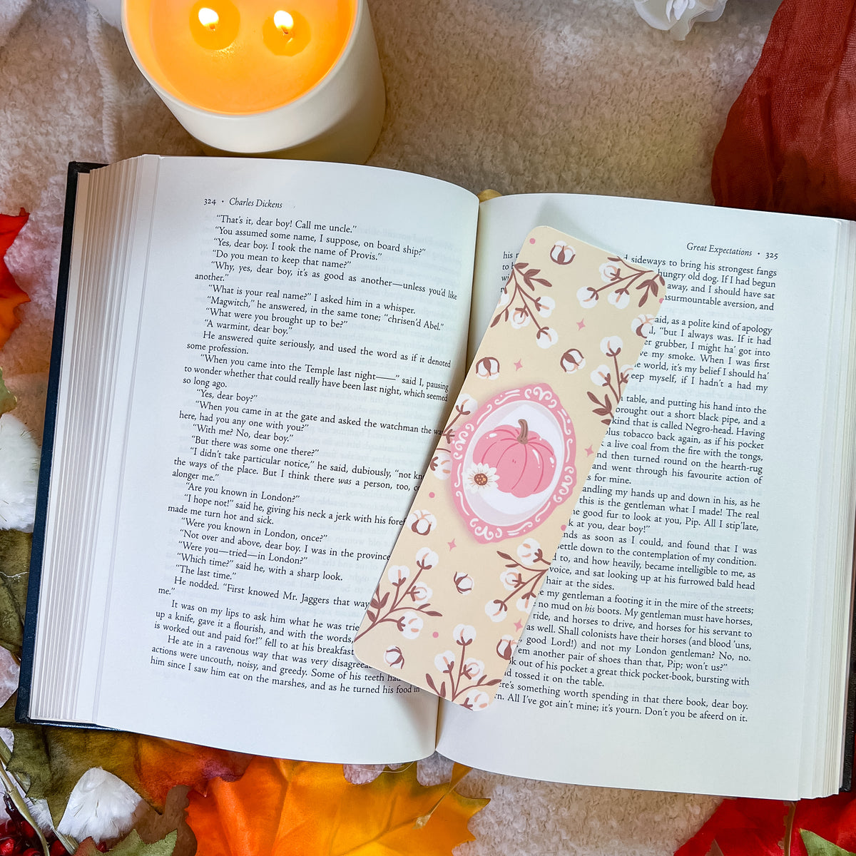 A decorative book bookmark with a pink pumpkin illustration and floral borders rests on an open book, surrounded by autumn leaves, a candle, and a red fabric background.