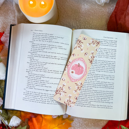 A decorative book bookmark with a pink pumpkin illustration and floral borders rests on an open book, surrounded by autumn leaves, a candle, and a red fabric background.