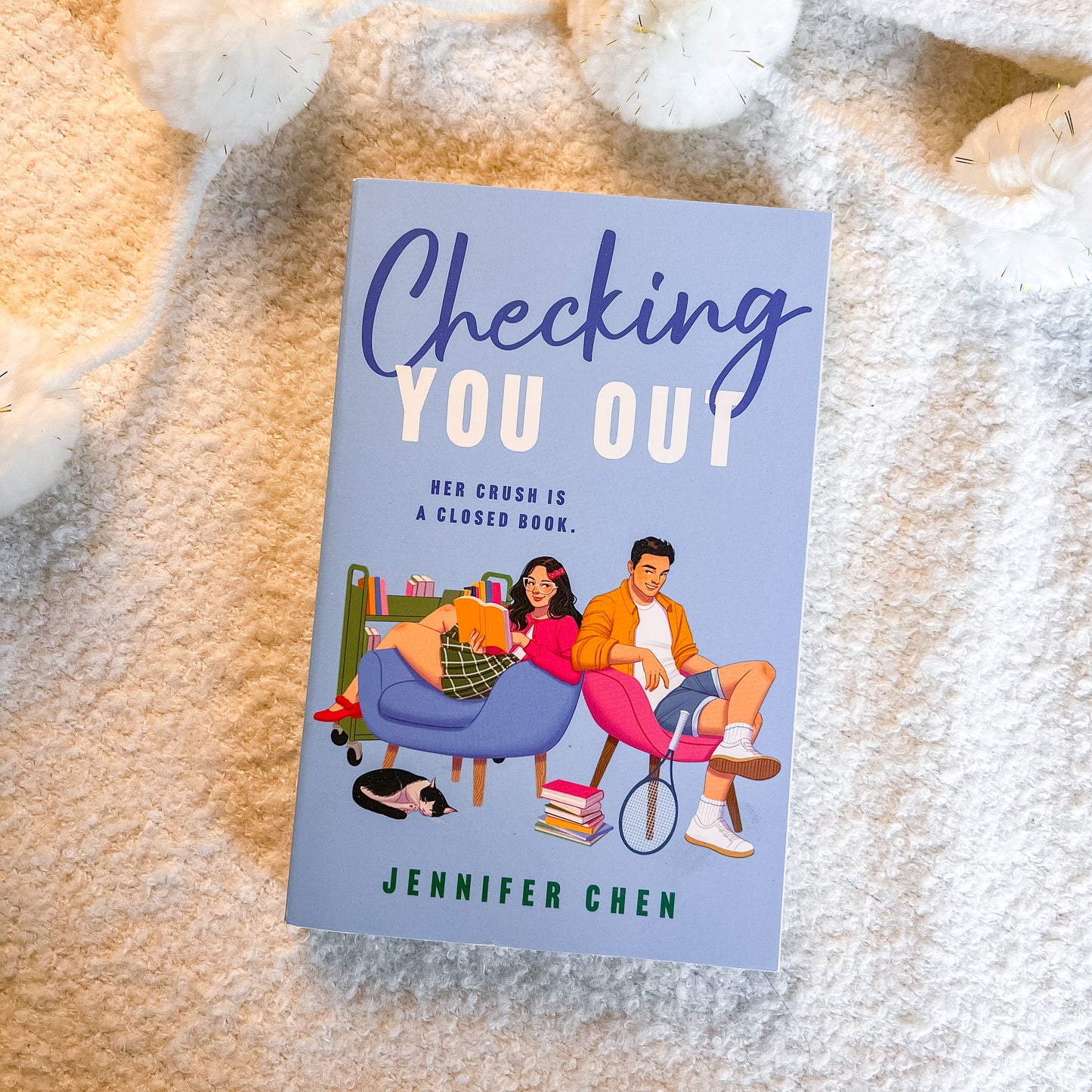 A book titled 'Checking You Out' by Jennifer Chen lies flat on a soft beige textured blanket, surrounded by white plush toys. The cover features a light blue background with an illustration of a young couple sitting on pink and blue chairs.
