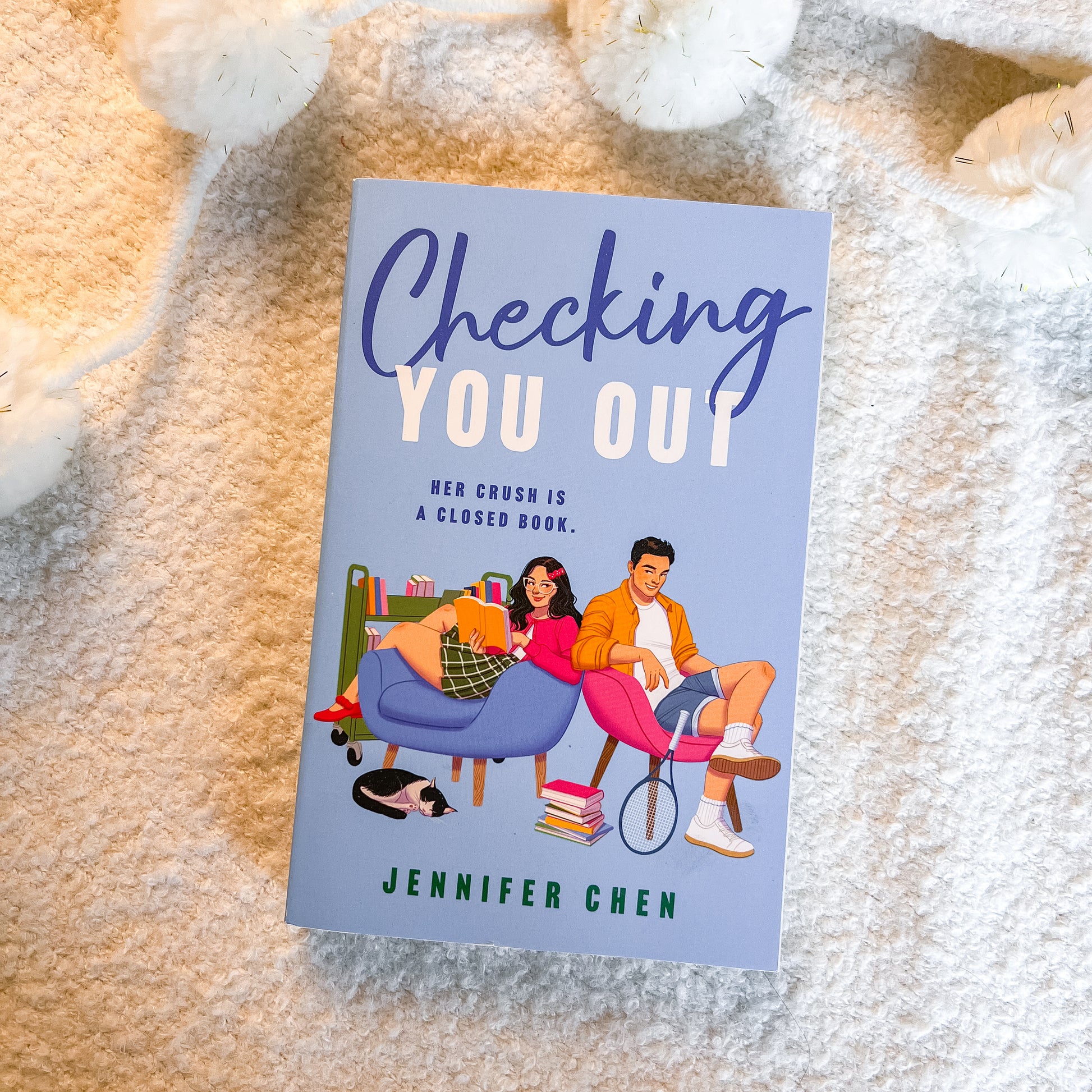 A book titled 'Checking You Out' by Jennifer Chen lies flat on a soft beige textured blanket, surrounded by white plush toys. The cover features a light blue background with an illustration of a young couple sitting on pink and blue chairs.