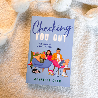 A book titled 'Checking You Out' by Jennifer Chen lies flat on a soft beige textured blanket, surrounded by white plush toys. The cover features a light blue background with an illustration of a young couple sitting on pink and blue chairs.