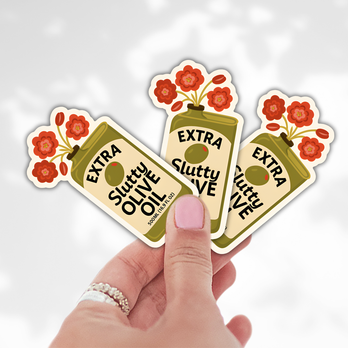 Hand holding three 'Extra Slutty Olive Oil' stickers with floral designs on a light gray background.