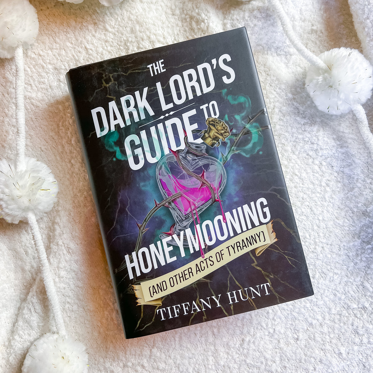 A book titled 'The Dark Lord's Guide to Honeymooning' by Tiffany Hunt, featuring a dark cover with a pink potion bottle illustration, placed on a white textured blanket with white pom-pom cords.