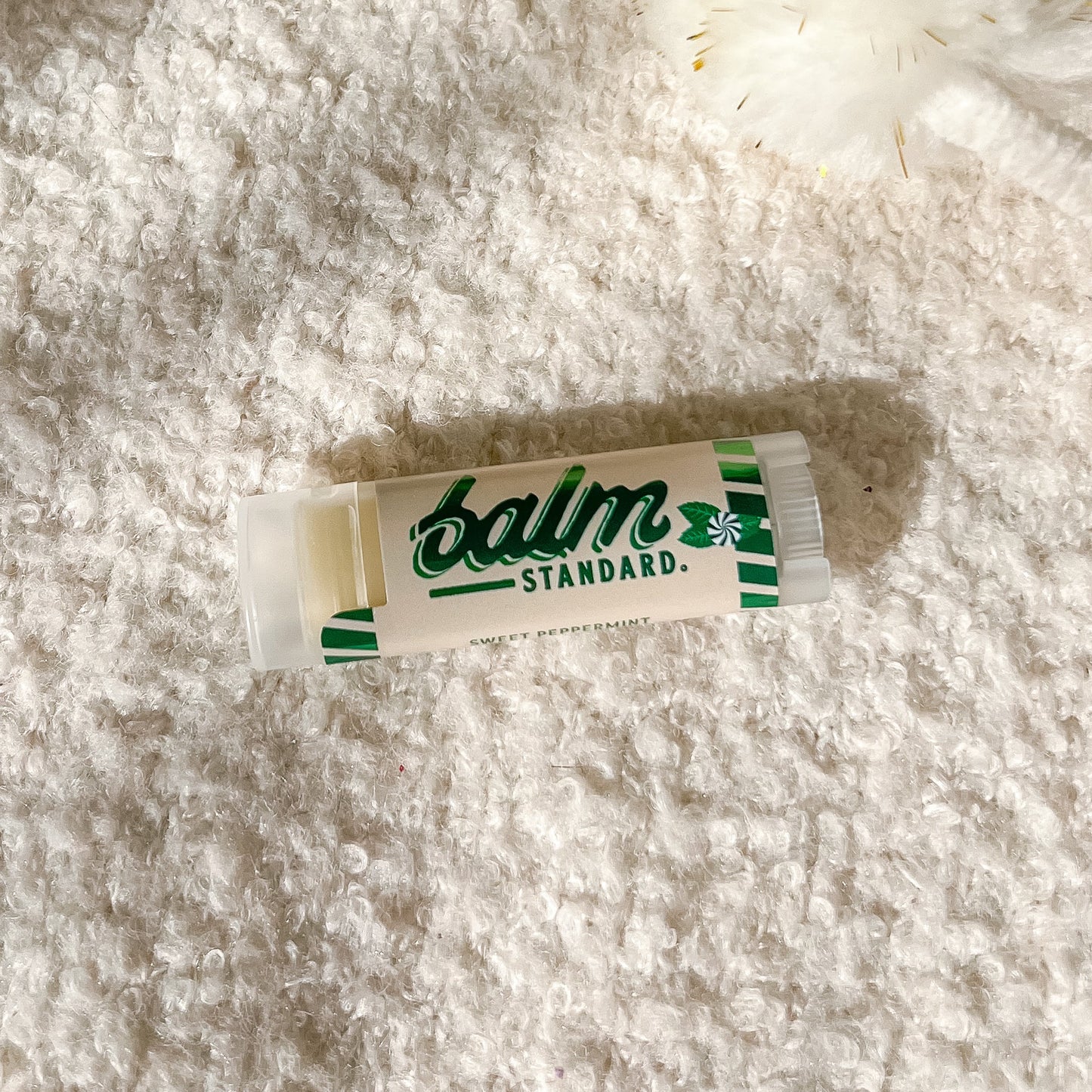 A tube of Balm Standard lip balm lies on a textured white fleece surface, with a white pom-pom accessory in the upper right corner.