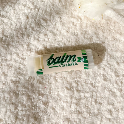 A tube of Balm Standard lip balm lies on a textured white fleece surface, with a white pom-pom accessory in the upper right corner.