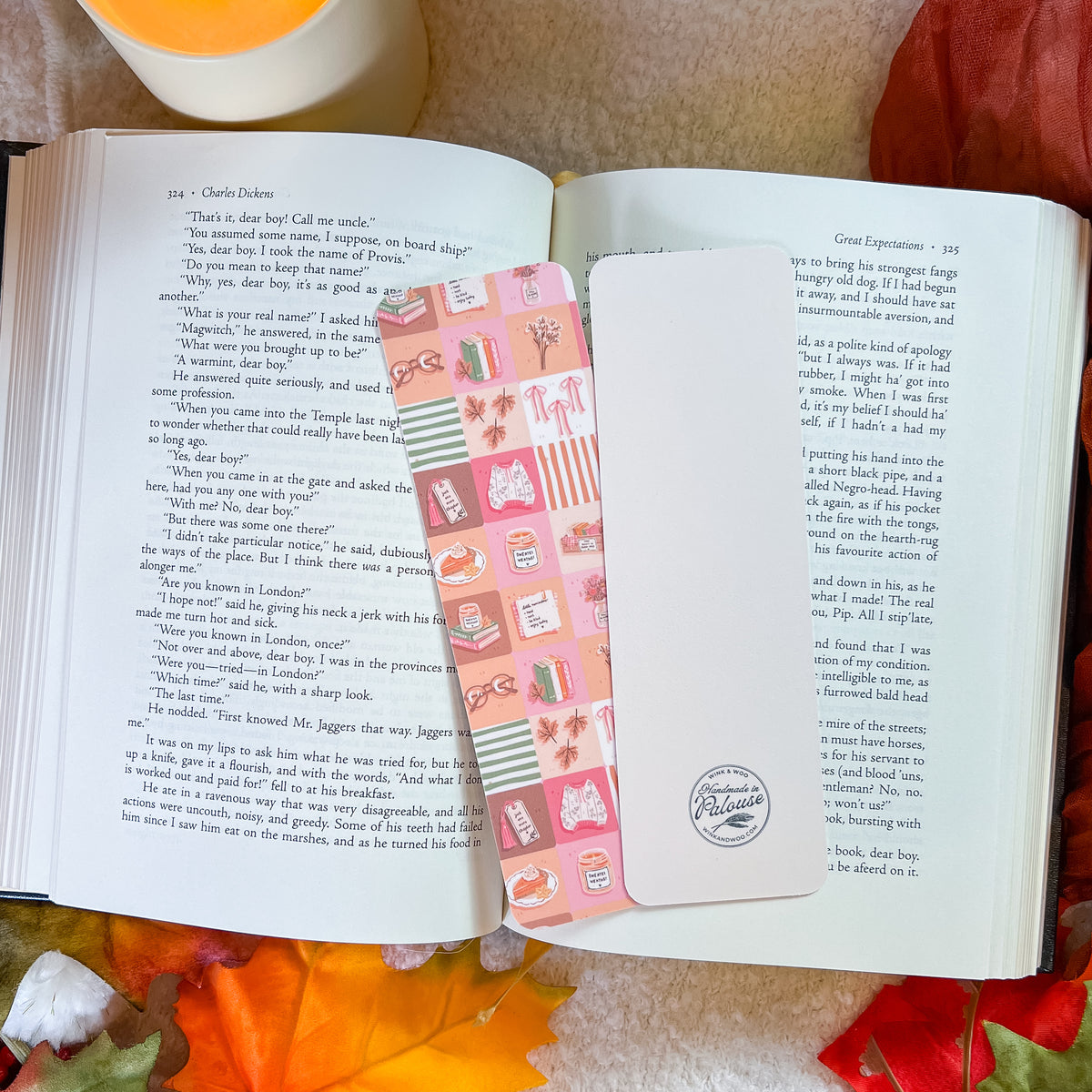 A pink and white patterned bookmark with cute illustrations is draped over an open book on a soft beige fabric background with autumn leaves.