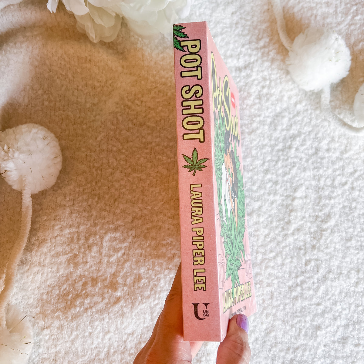 A hand holds a pink  book titled 'Pot Shot' by Laura Piper Lee. The book cover features a colorful illustration with green leaves and a woman.