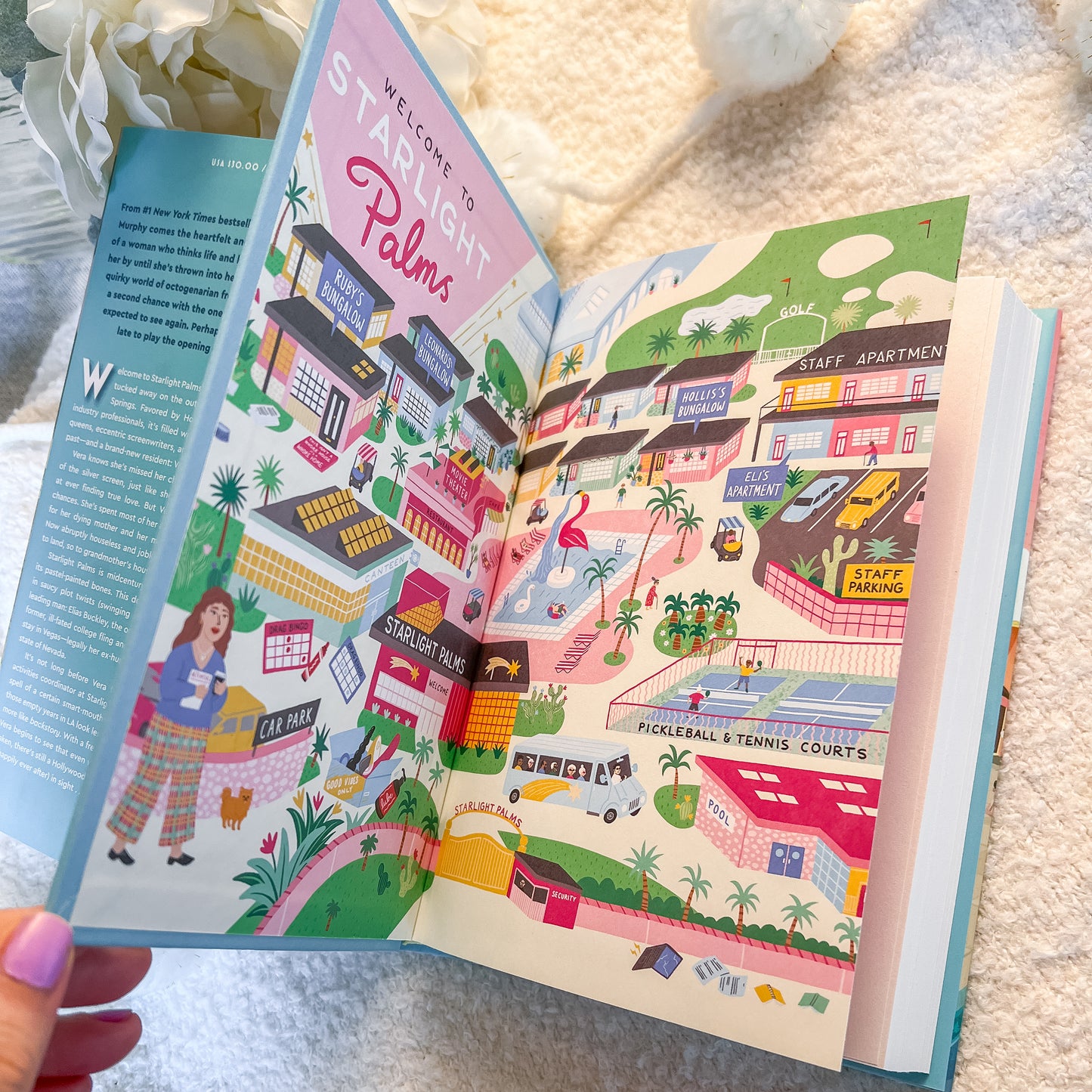 An open illustrated book spread showing colorful architectural city scenes with buildings, palm trees, people, and signage, photographed on a soft white textured blanket with a hand holding the left edge.
