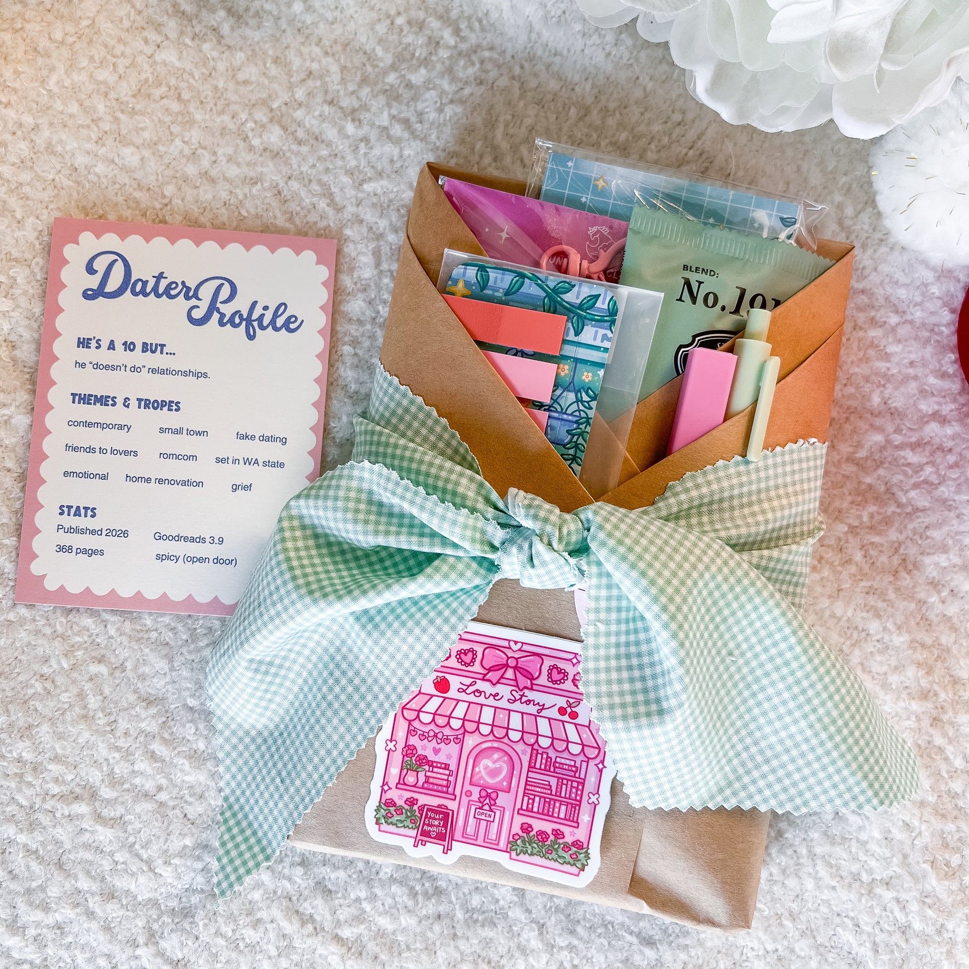 A gift set containing a 'Dater Profile' card, a brown kraft paper wrapped book filled with stationery items, and a mint green gingham ribbon tied in a bow with a pink house sticker. The set is arranged on a white textured blanket.