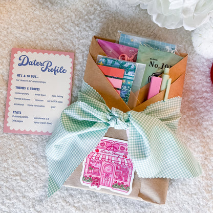 A gift set containing a 'Dater Profile' card, a brown kraft paper wrapped book filled with stationery items, and a mint green gingham ribbon tied in a bow with a pink house sticker. The set is arranged on a white textured blanket.