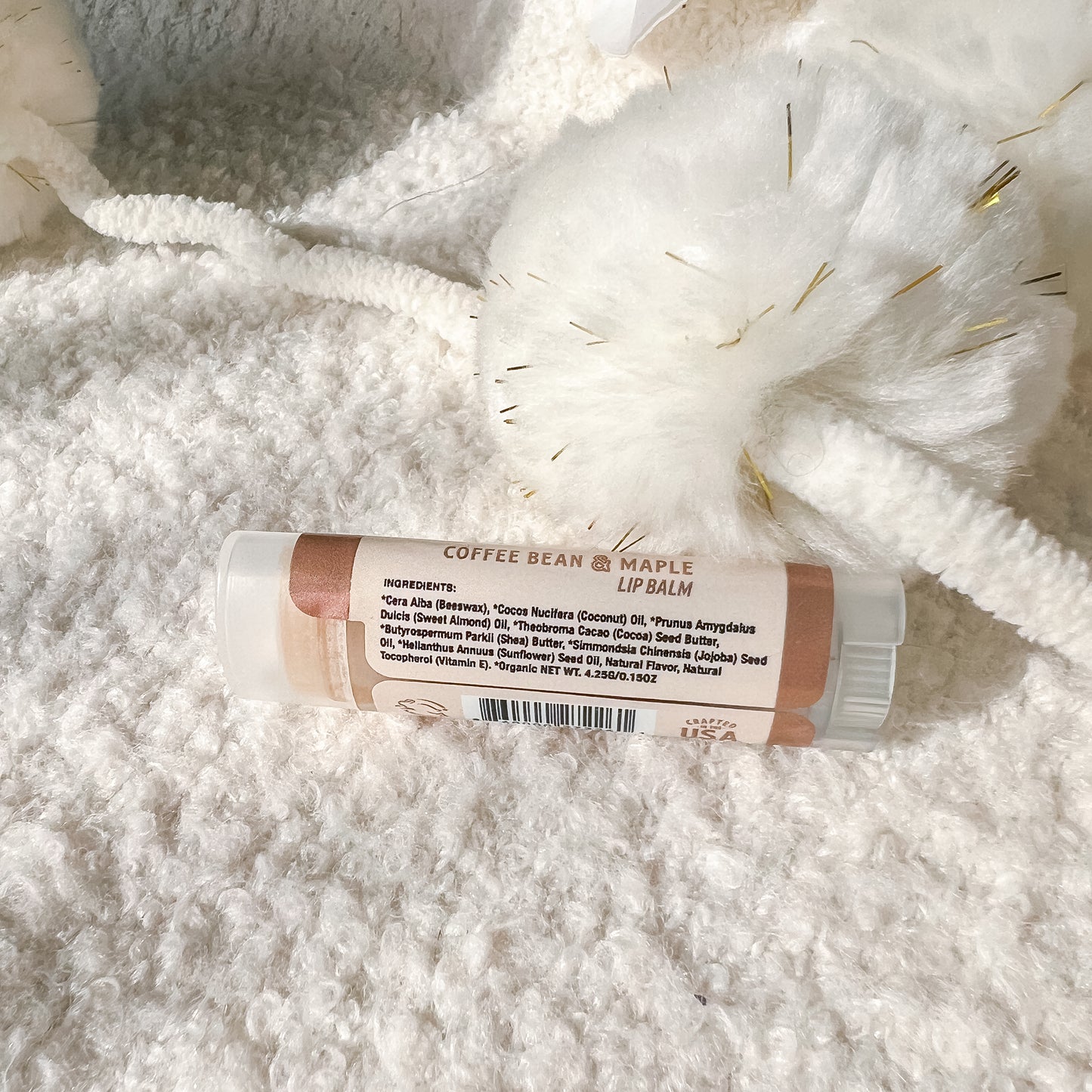 A tube of lip balm labeled 'COFFEE BEAN + MAPLE LIP BALM' resting on a soft white textured blanket with a white fluffy decorative piece behind it.
