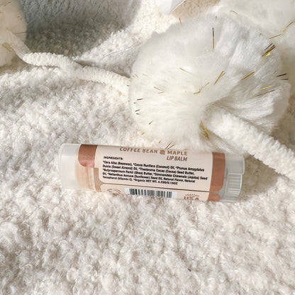 A tube of lip balm labeled 'COFFEE BEAN + MAPLE LIP BALM' resting on a soft white textured blanket with a white fluffy decorative piece behind it.
