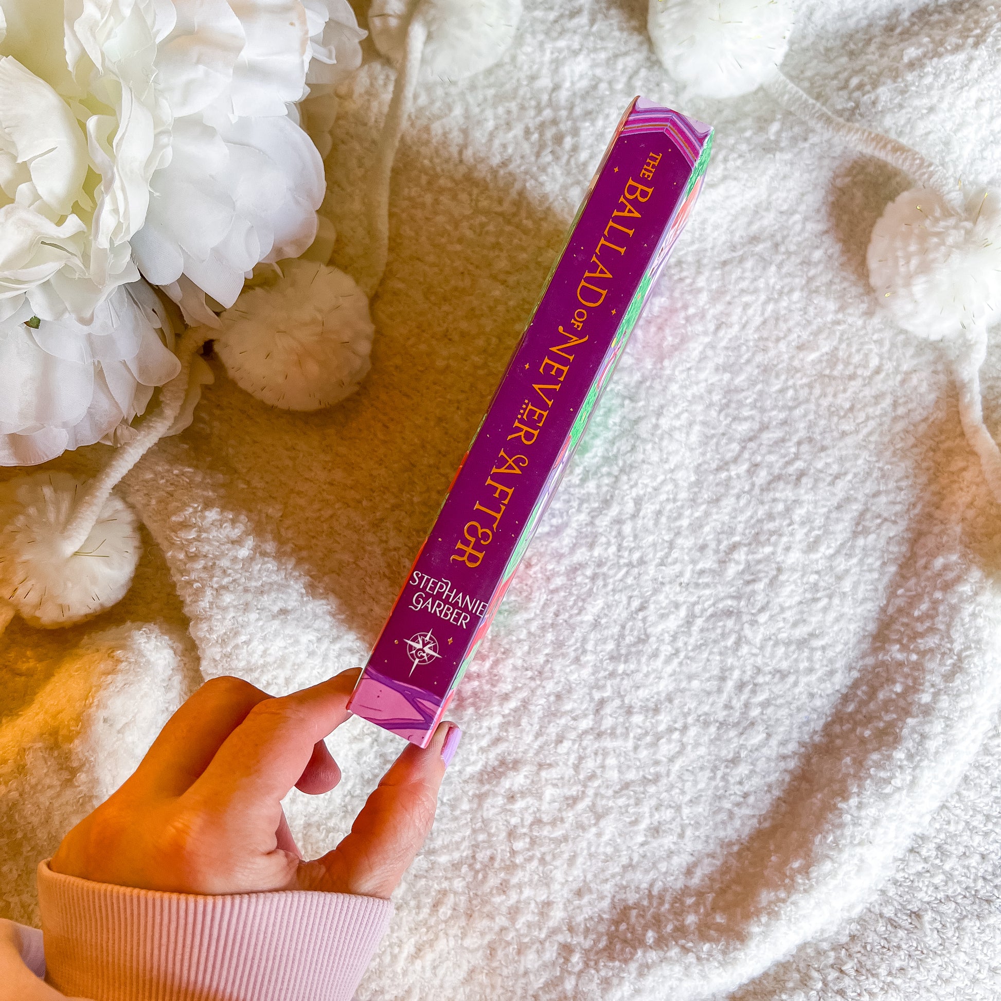 Hand holding a book titled 'The Ballad of Never After' by Stephanie Garber against a soft, blurred background with flowers.