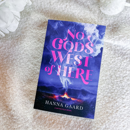 The book cover of 'No Gods West of Here' by Hanna Gaard, featuring a dark blue and purple smoky background with pink lettering, displayed on a soft beige textured blanket with white decorative flowers in the corners.