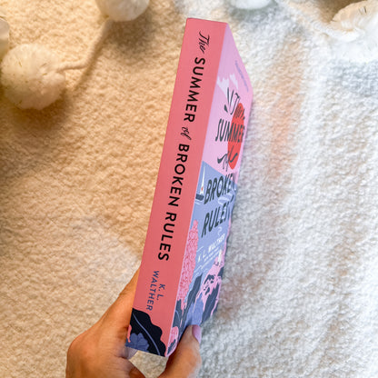 A hand holds a pink  book titled 'The Summer of Broken Rules' by K.L.