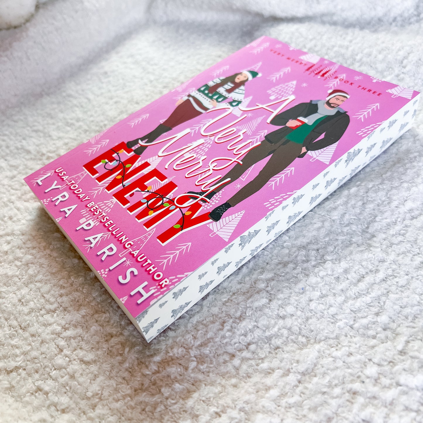 A book by Lyra Parish, featuring a bright pink cover with illustrations of a woman and a man in winter clothing, placed on a soft white textured blanket.