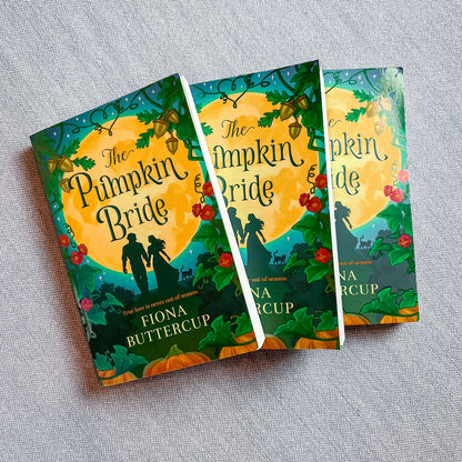 Three copies of the book 'The Pumpkin Bride' by Fiona Buttercup are arranged in a fan-like stack on a textured gray fabric background. The book cover features a vibrant autumn design with a large yellow moon, silhouettes of a couple and their dogs, pumpkins, and red berries on a green background.