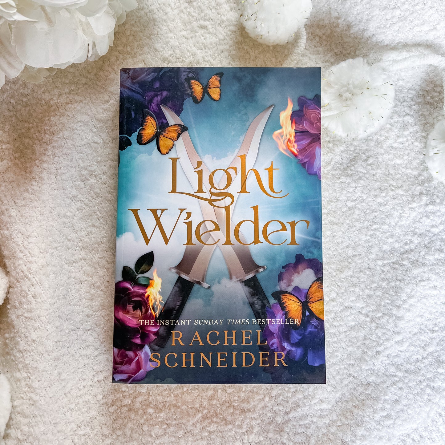 The book cover of 'Light Wielder' by Rachel Schneider is displayed flat on a textured white blanket, surrounded by decorative white flowers. The cover features a blue and purple fantasy background with crossed swords, glowing flames, and orange butterflies.
