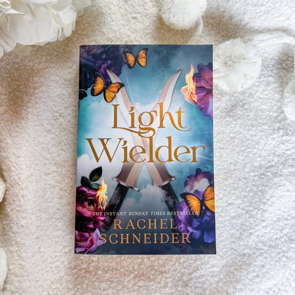 The book cover of 'Light Wielder' by Rachel Schneider is displayed flat on a textured white blanket, surrounded by decorative white flowers. The cover features a blue and purple fantasy background with crossed swords, glowing flames, and orange butterflies.