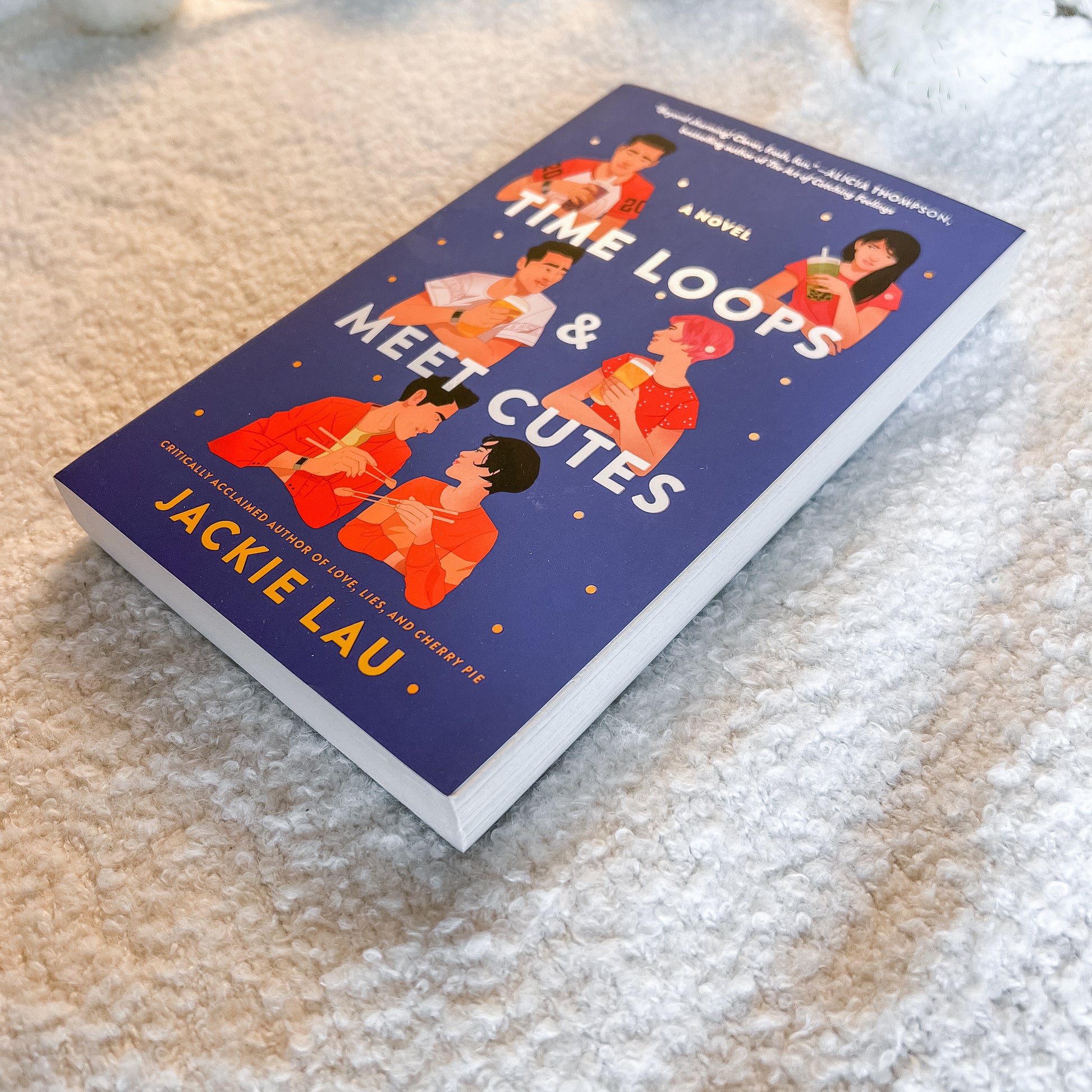 The book 'Time Loops & Meet Cutes' by Jackie Lau lies diagonally on a soft, textured beige carpet. The cover is dark blue with white and orange text and illustrations of characters in orange and pink outfits.