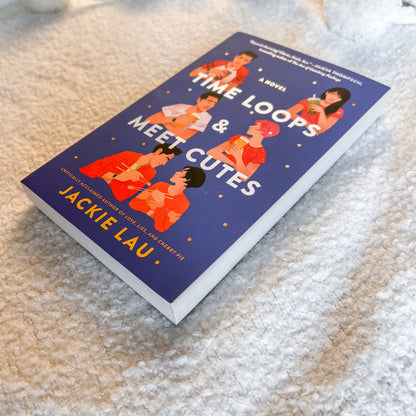 The book 'Time Loops & Meet Cutes' by Jackie Lau lies diagonally on a soft, textured beige carpet. The cover is dark blue with white and orange text and illustrations of characters in orange and pink outfits.