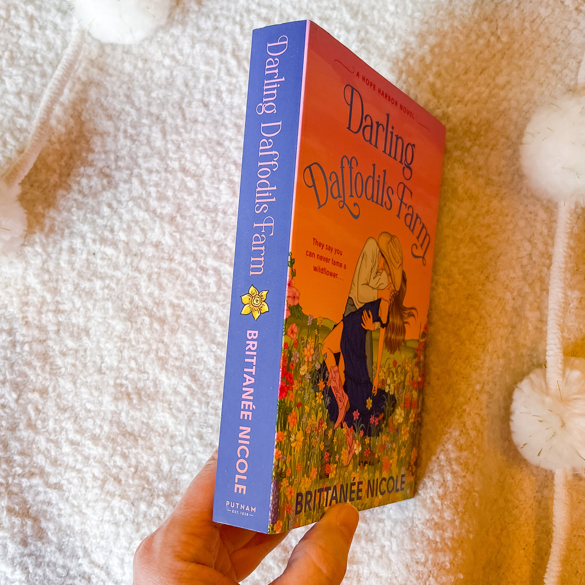 A hand holds a book titled 'Darling Delphids Farm' by Brittanie Nicole. The book has a blue spine and a warm orange cover featuring an illustration of a woman in a field.