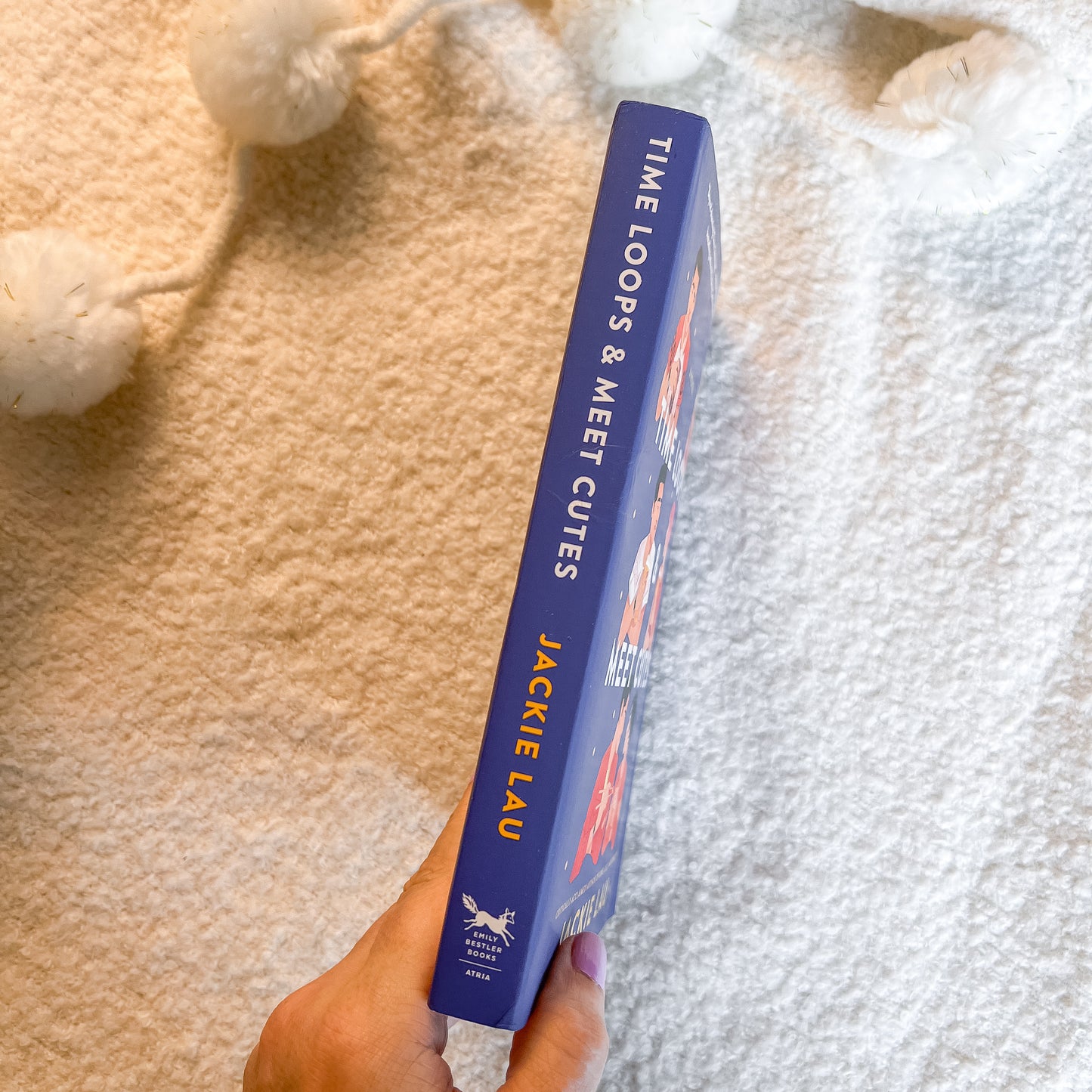 A hand holds a blue  book titled 'Time Loops & Meet Cutes' by Jacmie Lau, photographed from above on a textured cream blanket with white pom-pom garlands nearby.