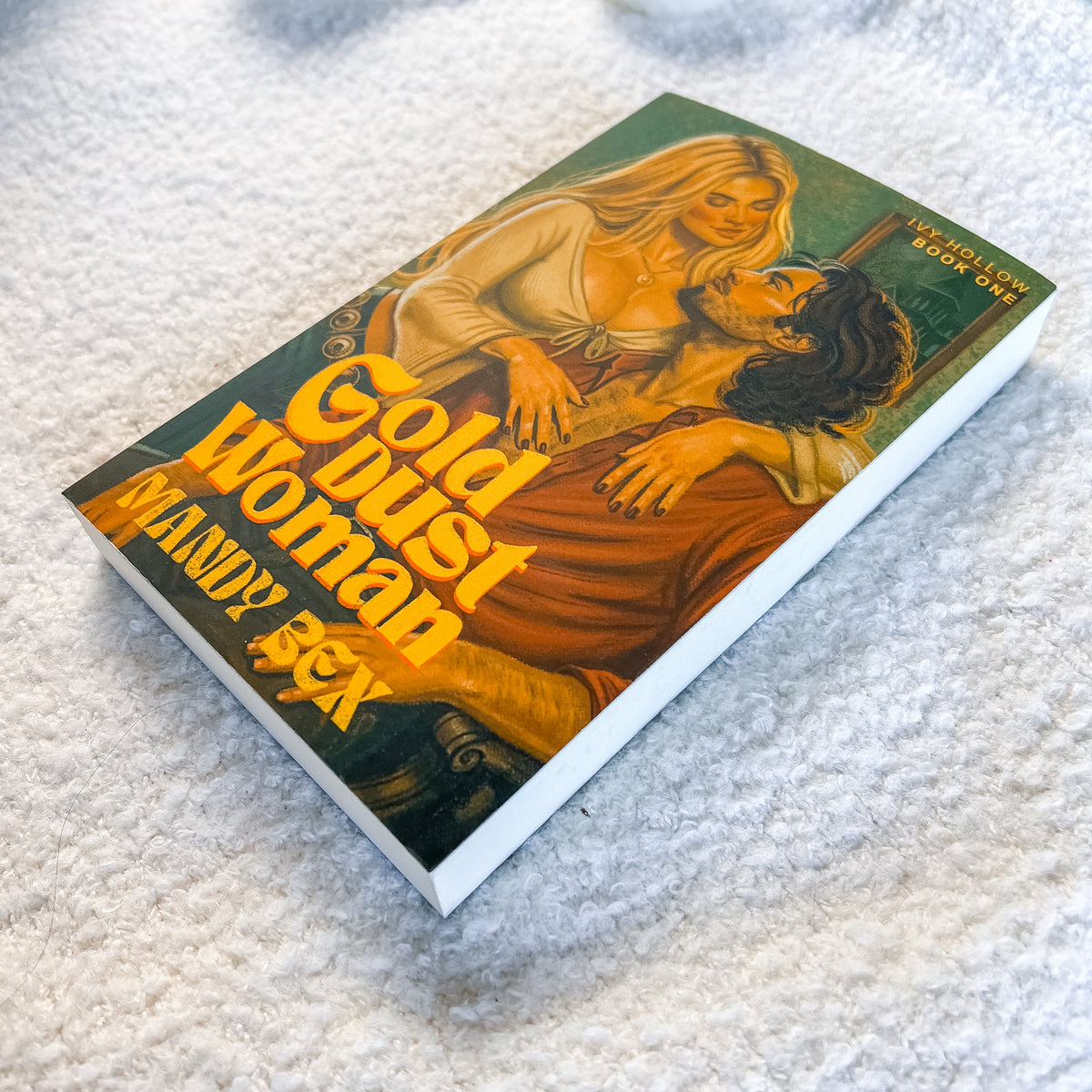 A paperback book titled 'Gold Dust Woman' by Mandy Bex, featuring a vintage-style illustration of a blonde woman and a dark-haired man on the cover, resting on a textured white fabric surface.