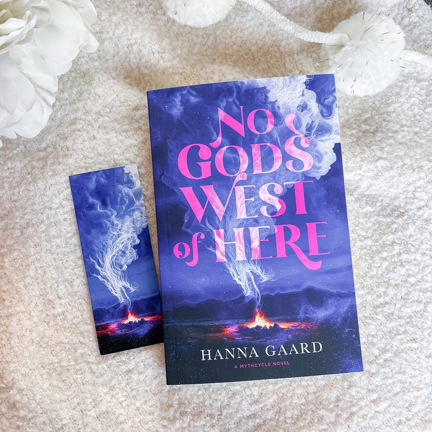 A book and a matching bookmark or smaller card featuring the cover art for 'No God's West of Here' by Hanna Gaard, styled on a soft beige textured blanket with white decorative flowers.
