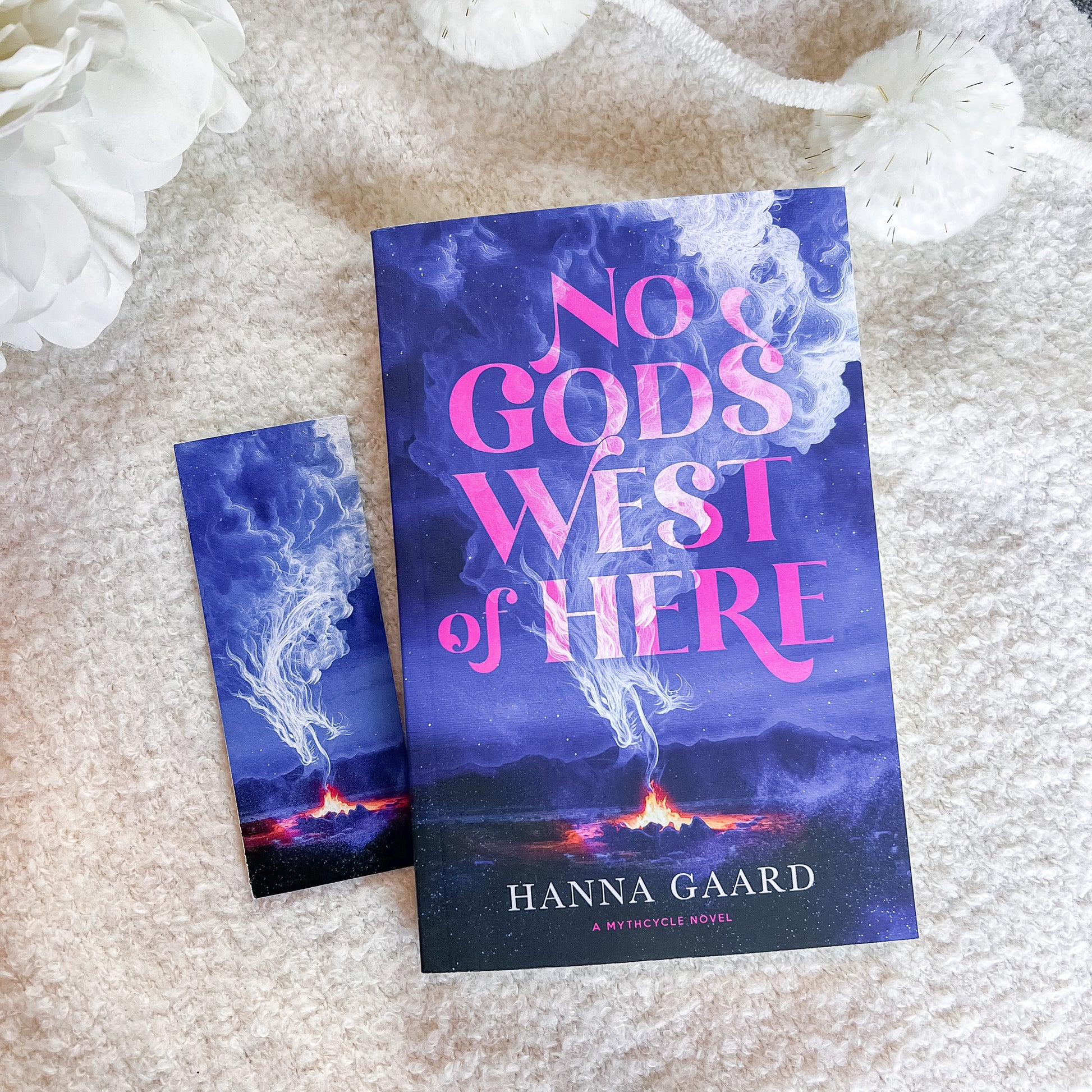 A book and a matching bookmark or smaller card featuring the cover art for 'No God's West of Here' by Hanna Gaard, styled on a soft beige textured blanket with white decorative flowers.