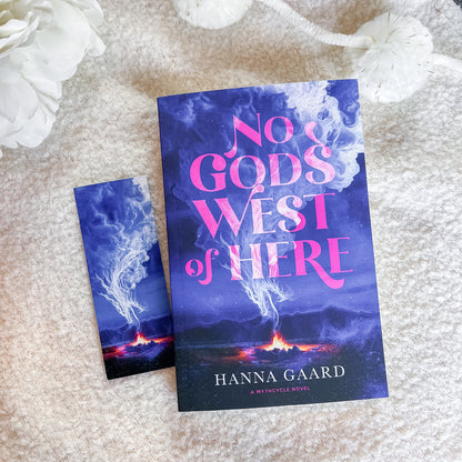 A book and a matching bookmark or smaller card featuring the cover art for 'No God's West of Here' by Hanna Gaard, styled on a soft beige textured blanket with white decorative flowers.