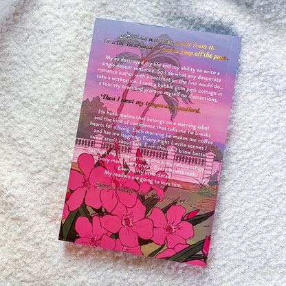 A book with a pink-to-purple gradient background, gold script text at the top, a black-and-white illustration of a grand building, and large pink flower illustrations along the bottom, photographed on a white textured fabric surface.