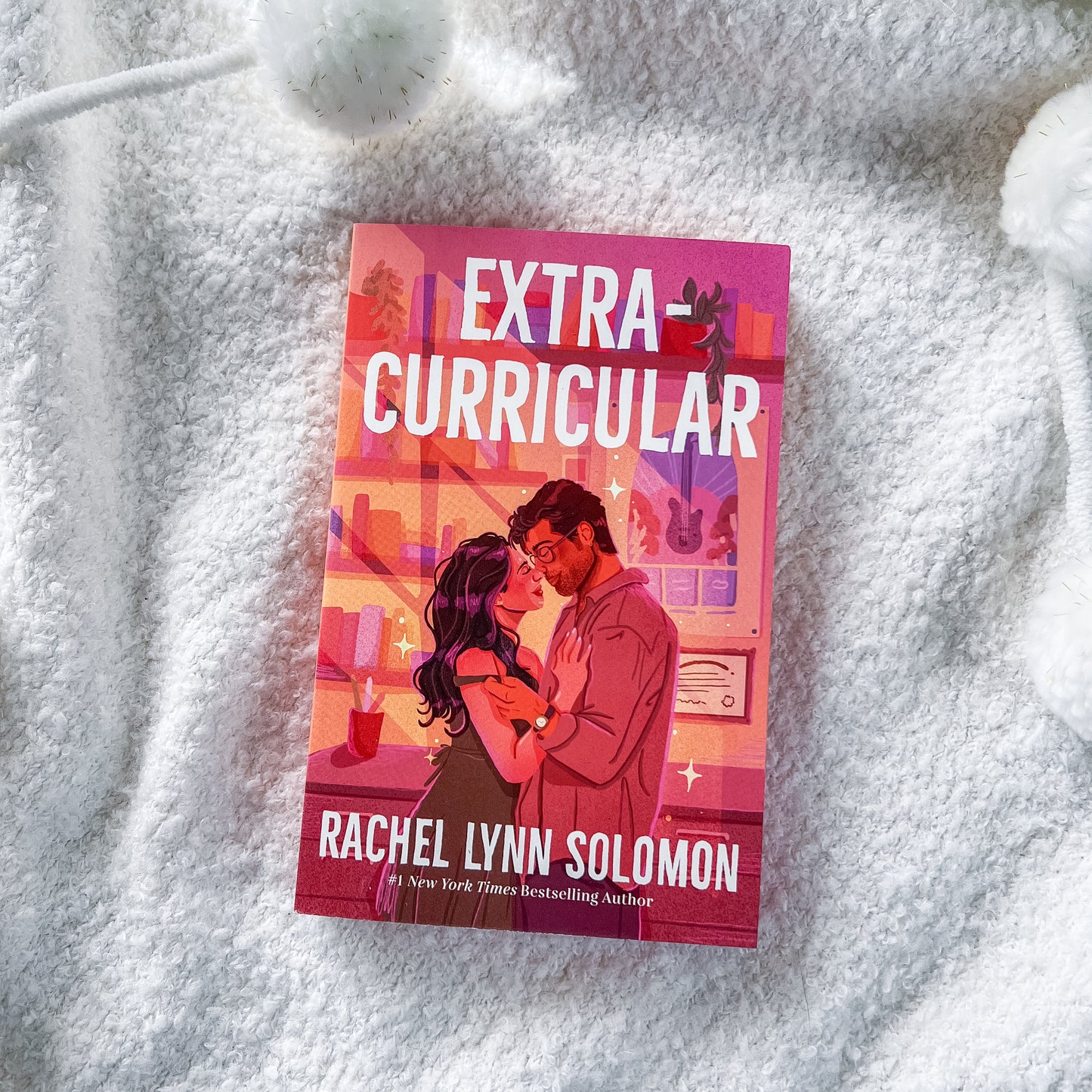 A book titled "Extra Curricular" by Rachel Lynn Solomon lies on a soft white textured blanket. The cover features a pink and orange-toned illustration of a couple embracing, with the title in large white letters at the top and the author's name at the bottom.