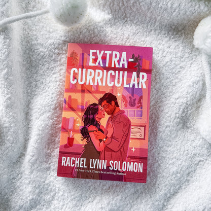 A book titled "Extra Curricular" by Rachel Lynn Solomon lies on a soft white textured blanket. The cover features a pink and orange-toned illustration of a couple embracing, with the title in large white letters at the top and the author's name at the bottom.