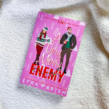 A book titled 'A Very Merry Enemy' by Lyra Parish, featuring a bright pink cover with an illustration of a couple in winter clothing, placed on a soft white textured blanket.