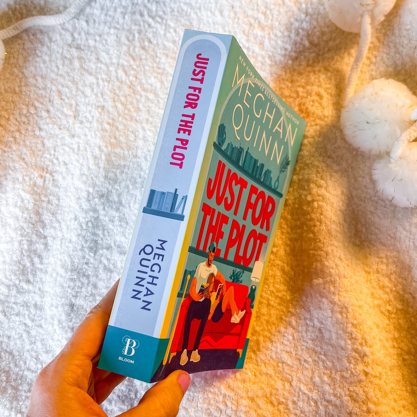 A hand holds a copy of the book 'Just For The Pilot' by Meghan Quinn, showing both the front cover and the spine. The book is photographed on a warm, textured white blanket with decorative white pom-poms in the background.