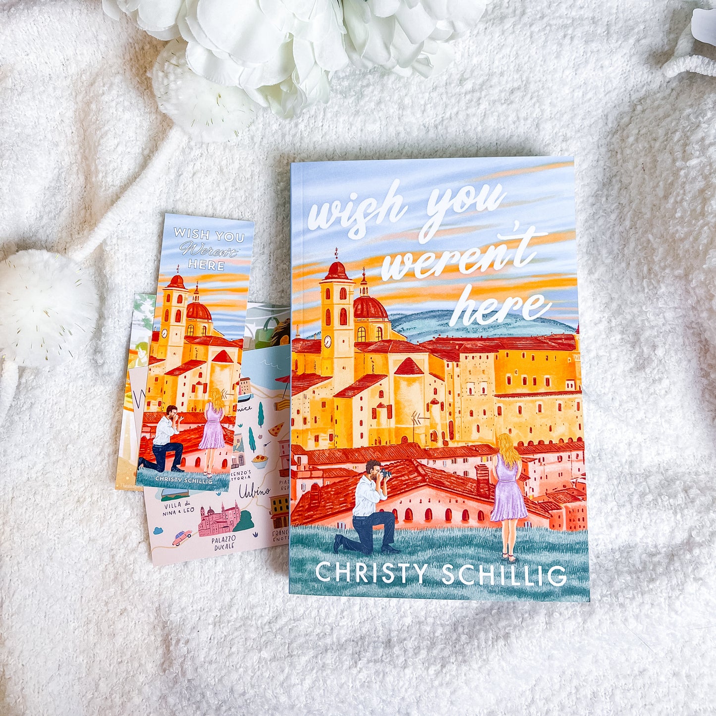 Two books titled 'wish you weren't here' by Christy Schillig on a textured white surface with white flowers.