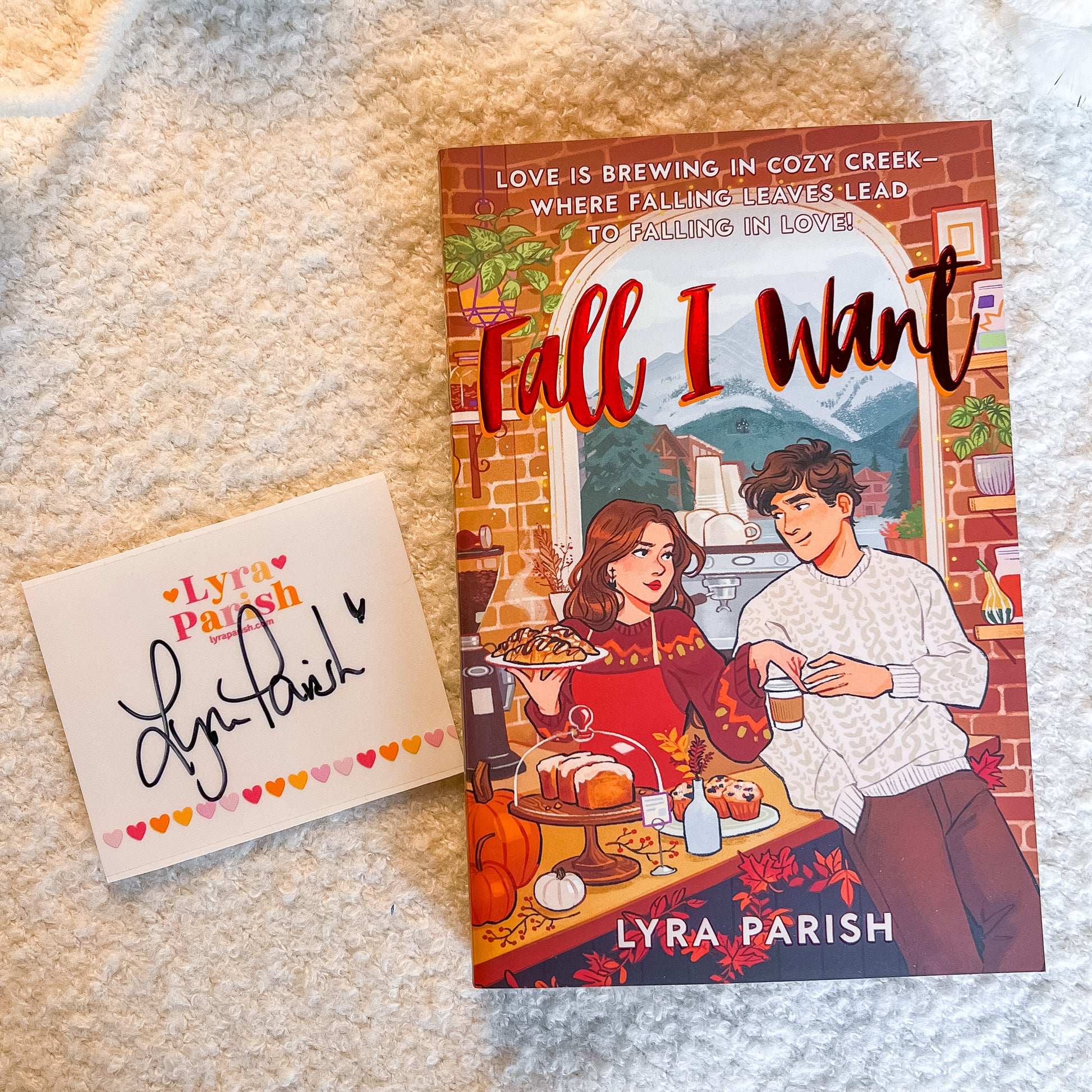 A book titled "Fall I Want" by Lyra Parish lies on a cream-colored textured blanket next to a matching white card with a black signature and small heart border. The book cover features an illustrated couple in a cozy kitchen scene with autumn decorations.