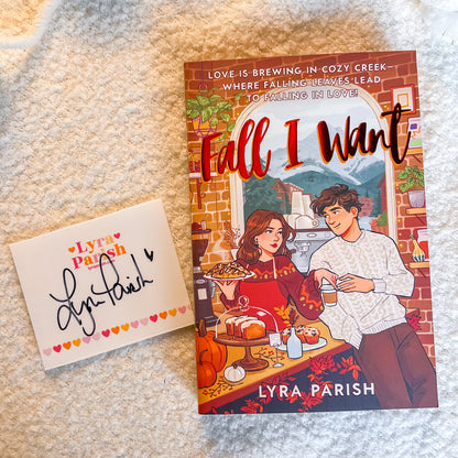 A book titled "Fall I Want" by Lyra Parish lies on a cream-colored textured blanket next to a matching white card with a black signature and small heart border. The book cover features an illustrated couple in a cozy kitchen scene with autumn decorations.