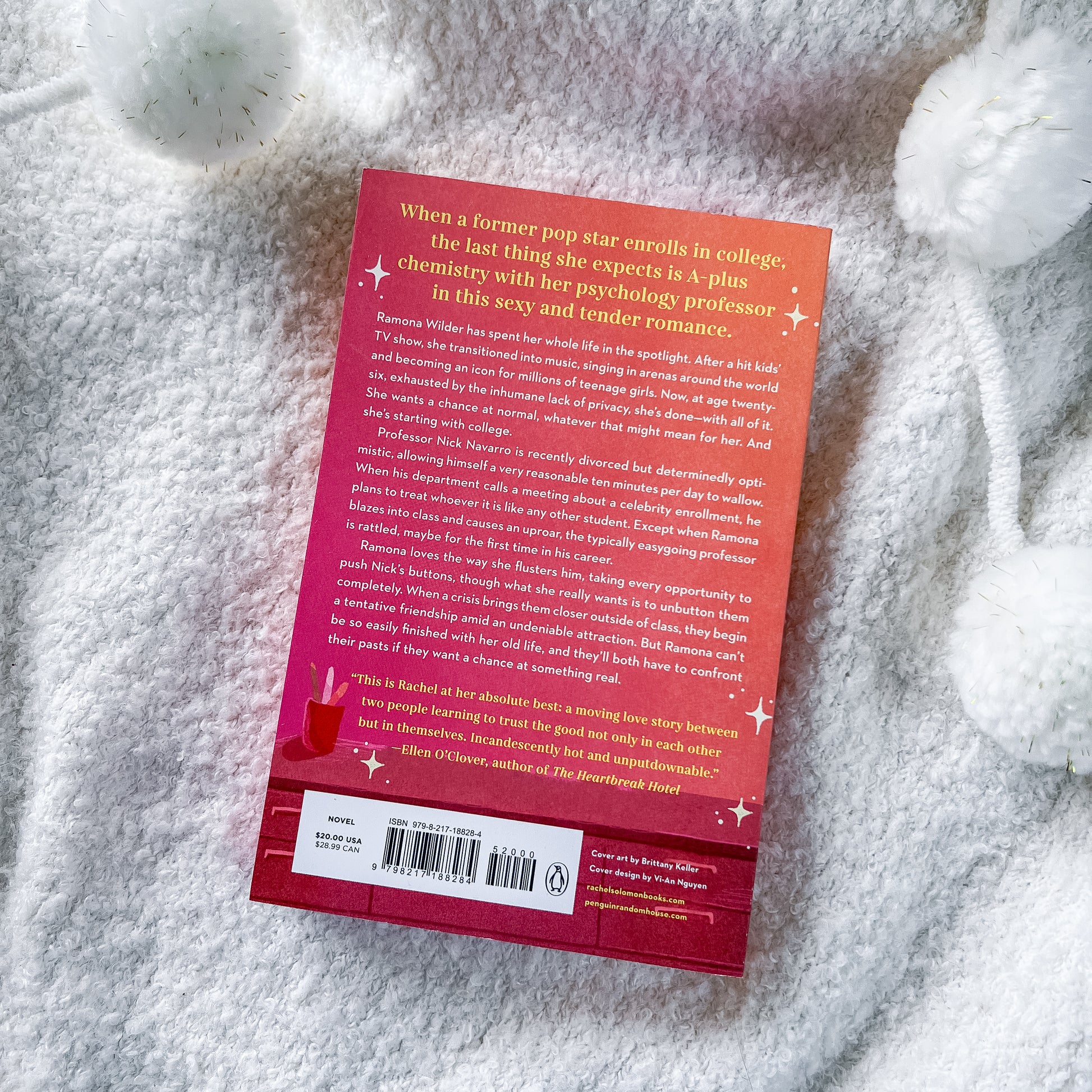 The back cover of the book displayed flat on a textured white blanket with decorative white flowers. The cover features a vibrant red-to-pink gradient with white text and small star accents.
