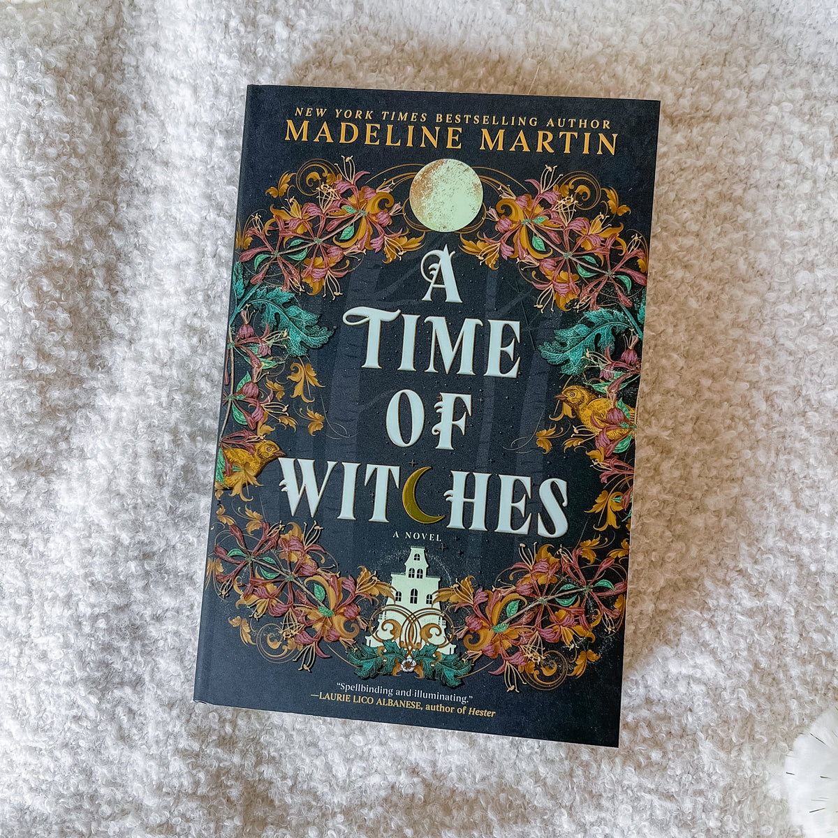 The book cover of 'A Time of Witches' by Madeline Martin, featuring a dark background with intricate floral patterns and a full moon, photographed flat on a textured white fabric surface.