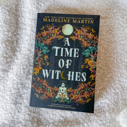 The book cover of 'A Time of Witches' by Madeline Martin, featuring a dark background with intricate floral patterns and a full moon, photographed flat on a textured white fabric surface.