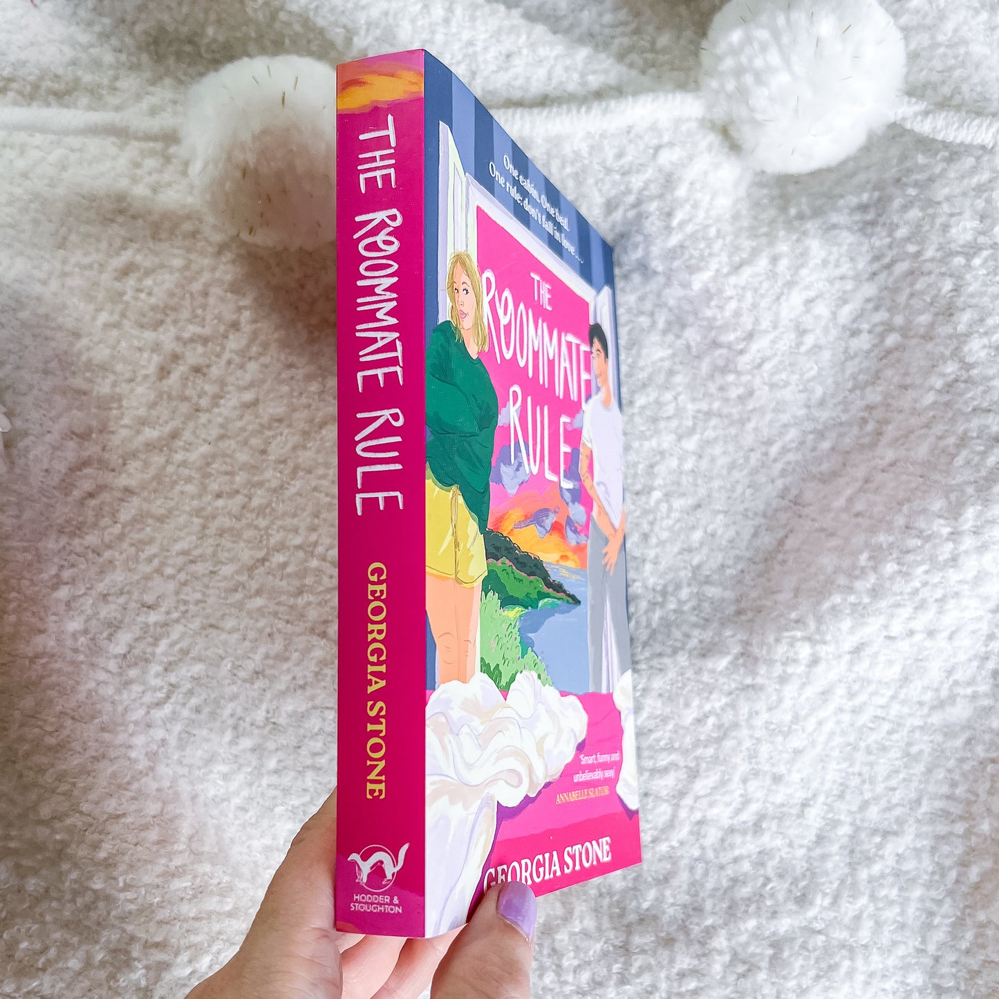 A hand holds a copy of the book 'The Roommate Rule' by Georgia Stone. The book cover features a colorful illustration of a woman in a green jacket and yellow skirt, and the spine is bright pink with the title and author name in gold.