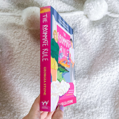 A hand holds a copy of the book 'The Roommate Rule' by Georgia Stone. The book cover features a colorful illustration of a woman in a green jacket and yellow skirt, and the spine is bright pink with the title and author name in gold.