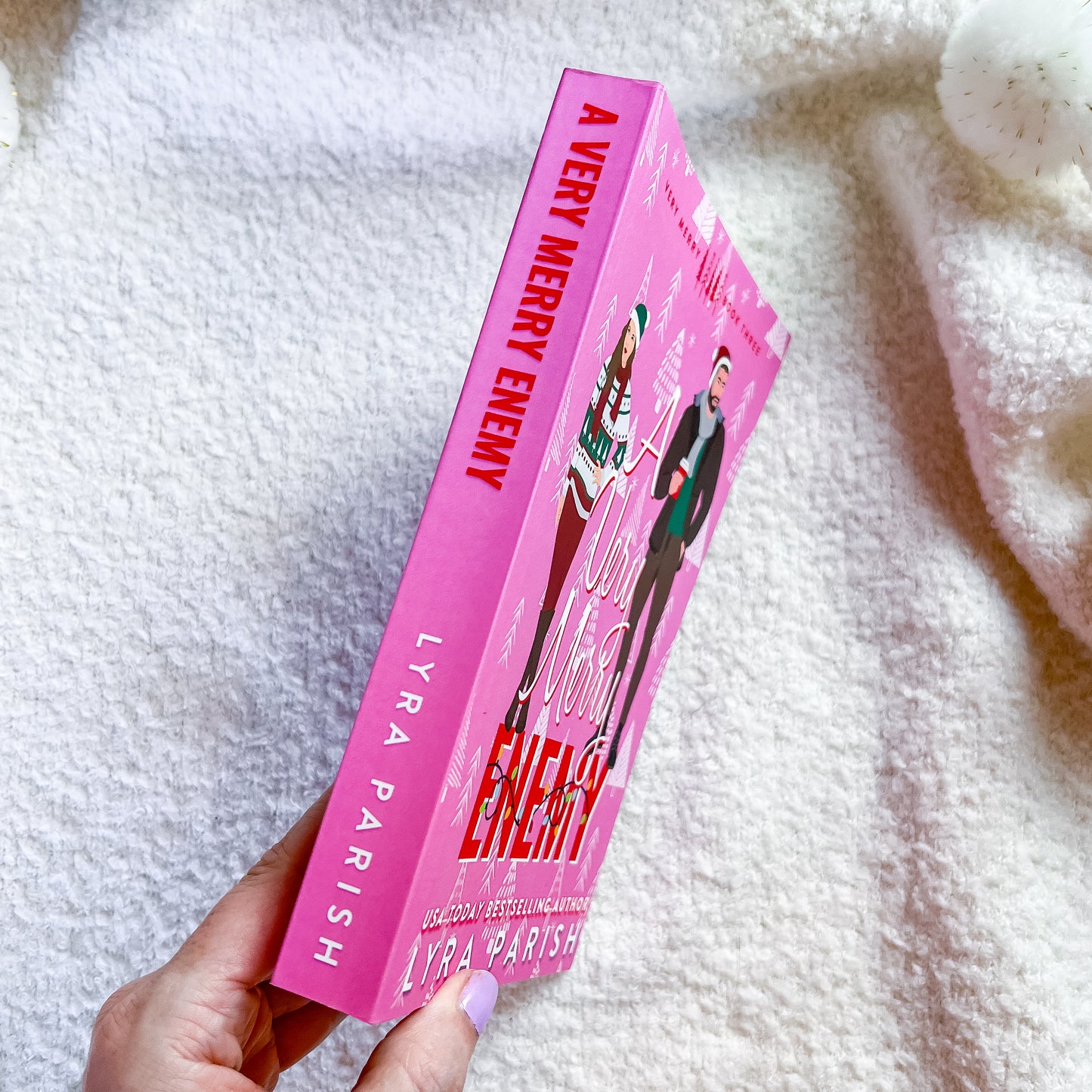 A hand holds a pink copy of the book 'A Very Merry Enemy' by Lyra Parish, featuring illustrations of characters on the cover, set against a soft white textured blanket background.